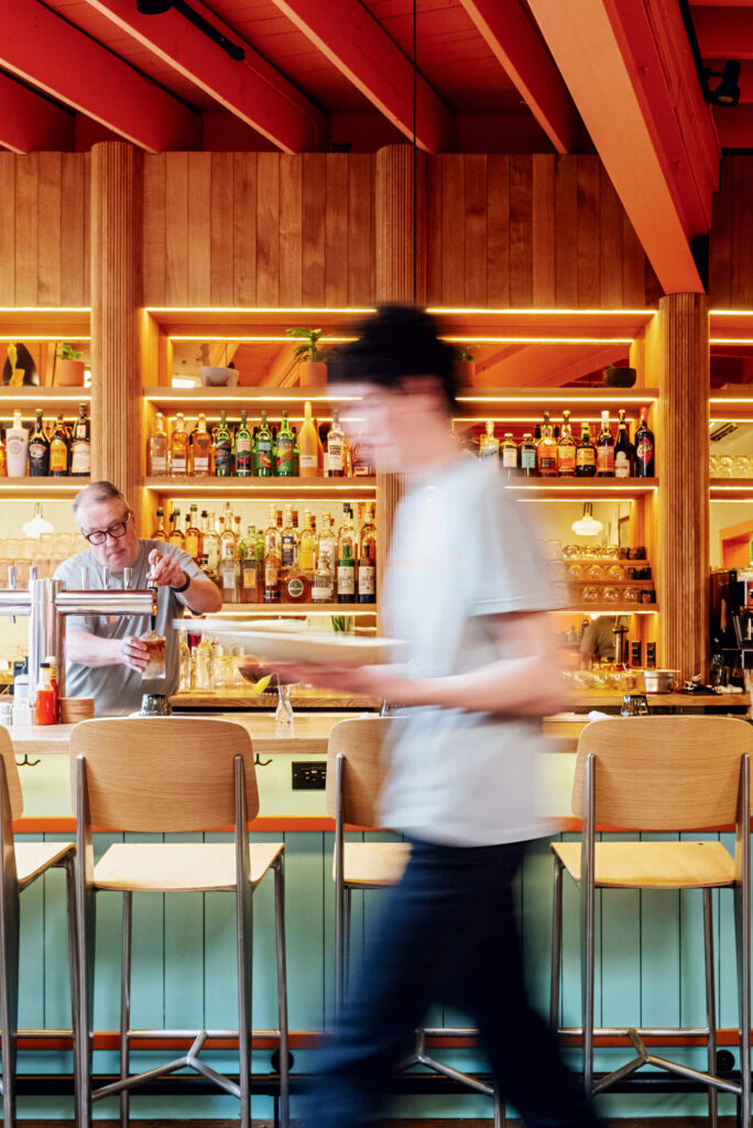 Bartender prepares a drink behind a well-stocked bar as a person walks past empty bar stools.