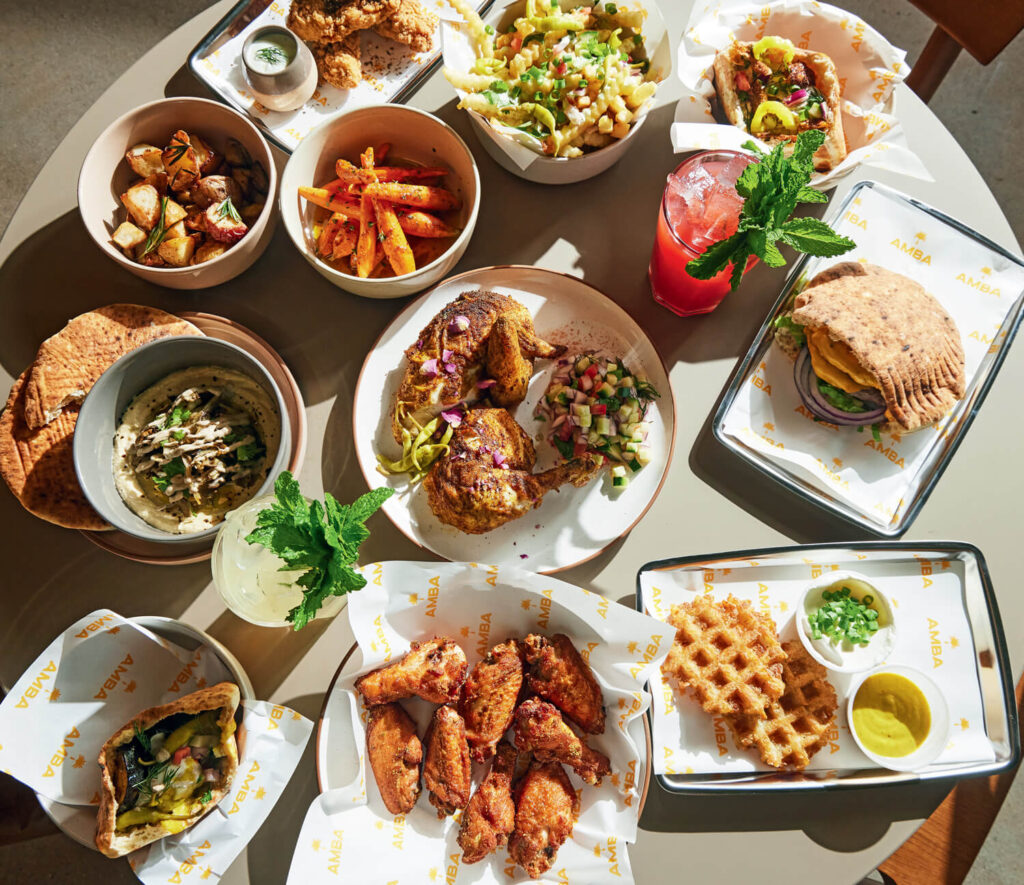 A table filled with assorted Mediterranean dishes, fried chicken, waffle fries, salads, and colorful drinks.