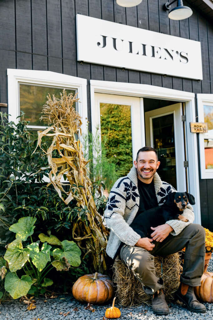 Man sitting on hay bale with a black dog in front of a shop called JULIEN'S, surrounded by autumn decor.