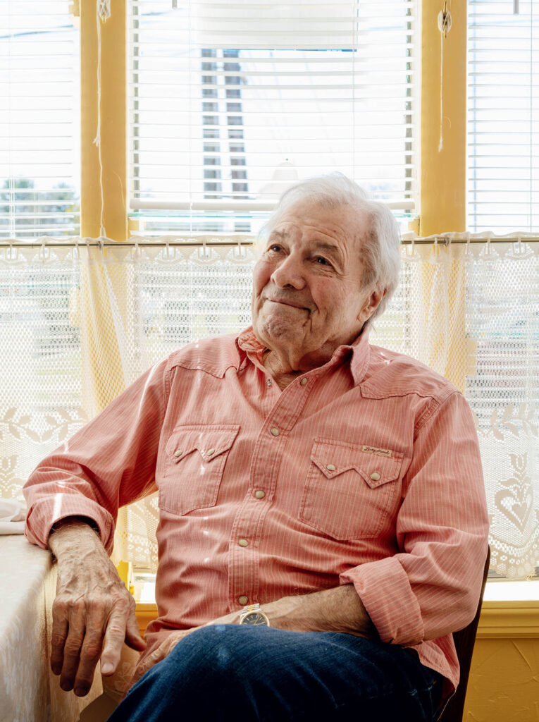 Jacques Pépin sits by a window with lace curtains, smiling thoughtfully.