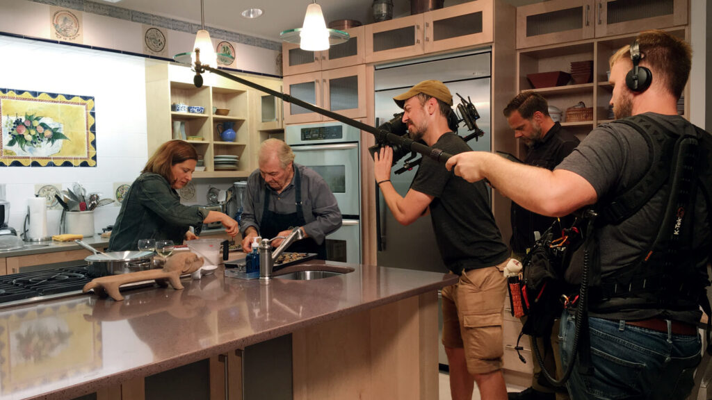 Two people cook in a kitchen while a film crew records them with a camera and boom microphone.