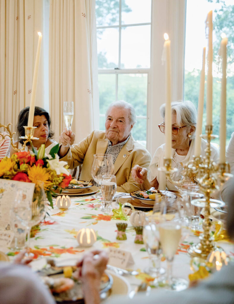 Elderly people sitting at a decorated dinner table, raising glasses in a toast, with candles and flowers.
