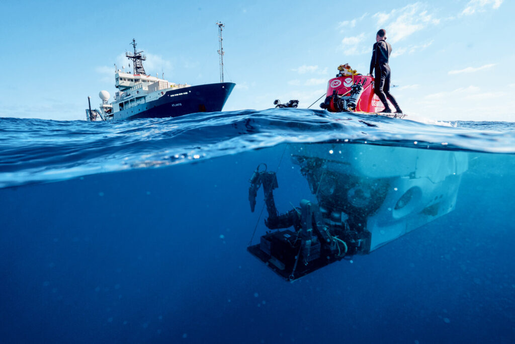 A diver on a small boat floats above a submersible underwater, with a research ship in the background.