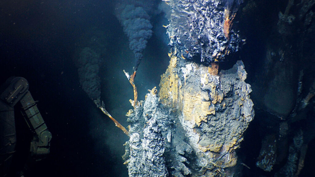 A hydrothermal vent on the seafloor emits dark plumes, with a robotic arm visible on the left.