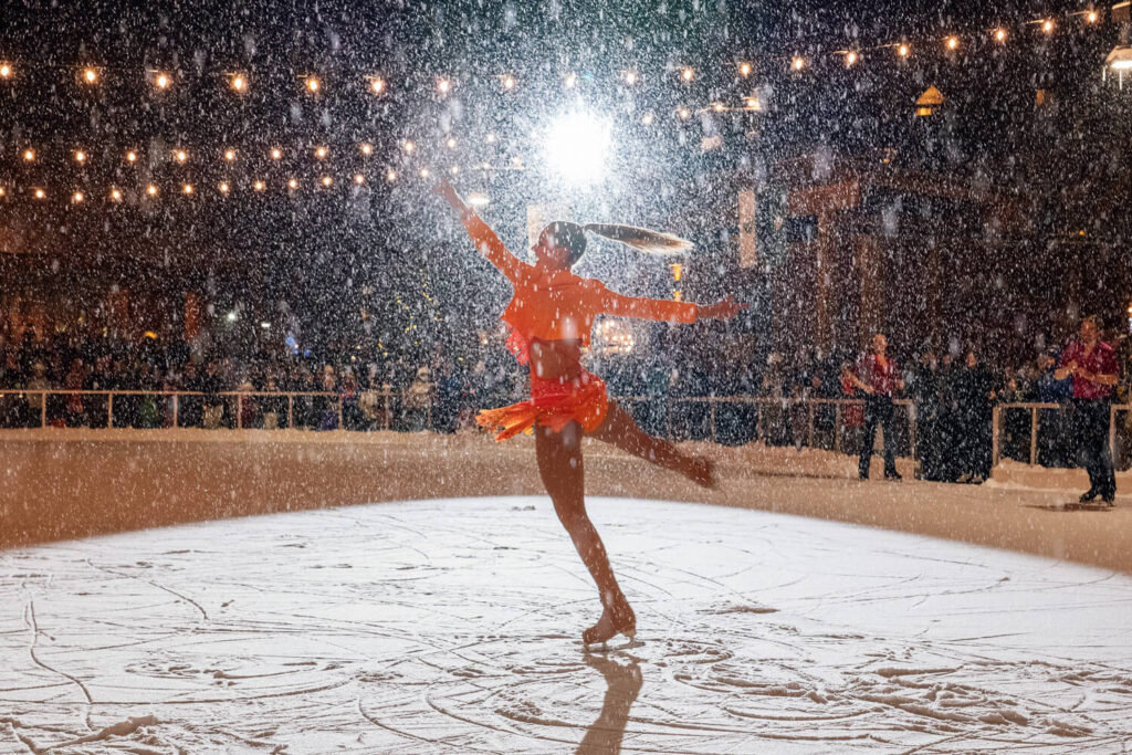 Figure skater in an orange outfit performs under falling snow with bright lights and a cheering crowd.