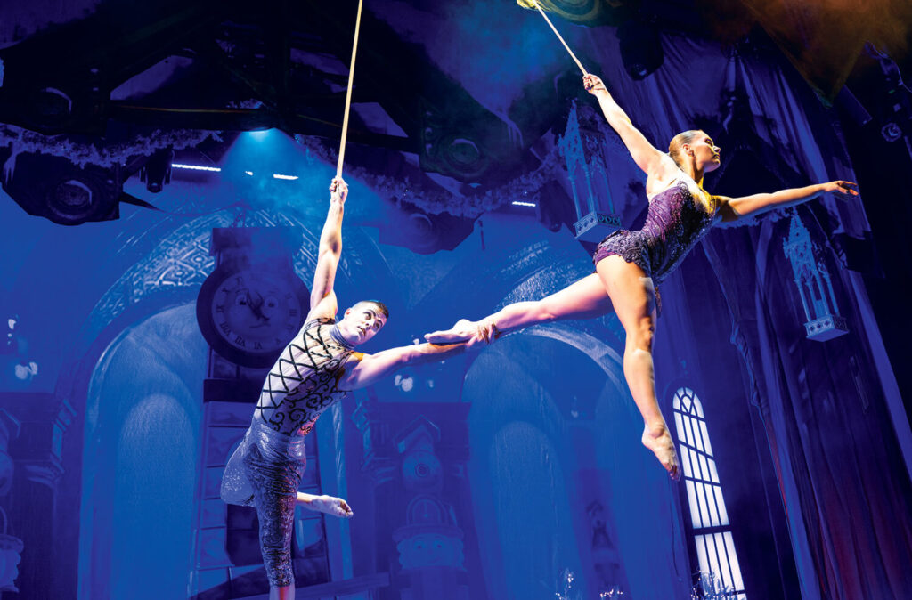 Two acrobats perform a balancing aerial act on ropes on a brightly lit stage with a blue and purple backdrop.