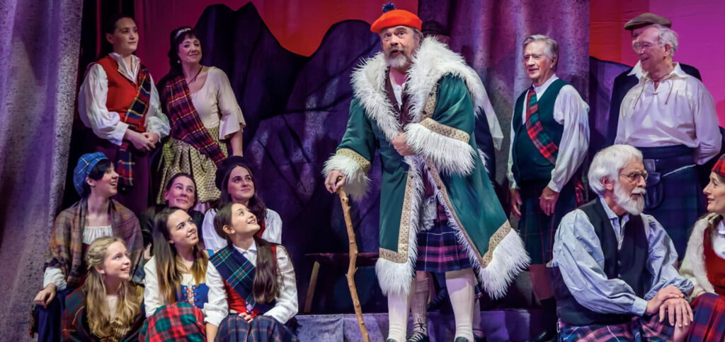 A man in a green fur-trimmed coat and red beret performs on stage with others in plaid costumes.