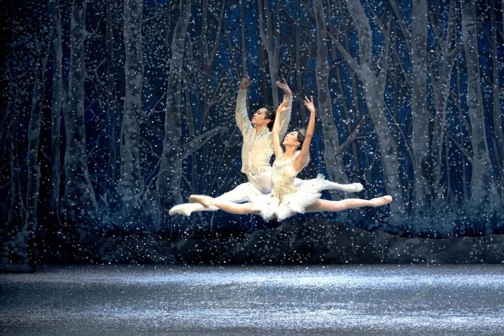 Two ballet dancers leap gracefully on stage, surrounded by falling snow and a wintry forest backdrop.