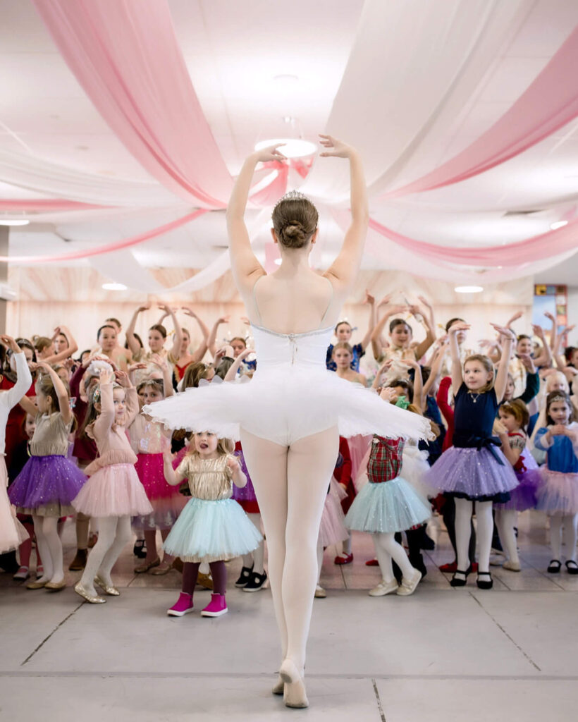 Ballet dancer on stage leads a group of children in colorful tutus, all posing with arms raised.