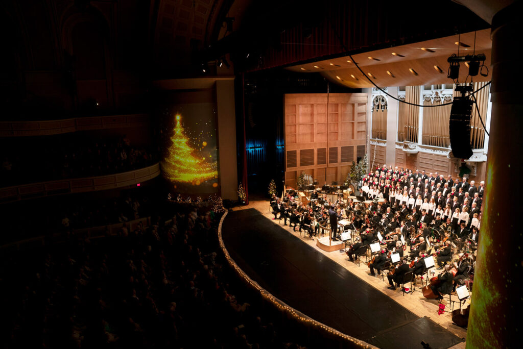 A symphony orchestra and choir perform onstage with a glowing Christmas tree projected on the wall.