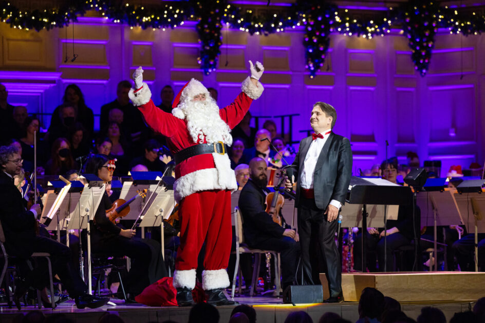 Santa Claus and a conductor stand onstage with an orchestra, festive lights and decorations in the background.