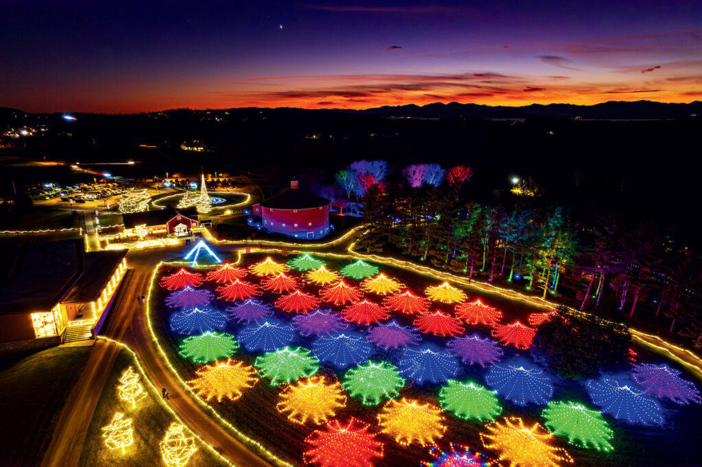 Winter Lights at Shelburne Museum: A Vermont Holiday Light Display. A field displays colorful holiday lights in patterns at sunset, with trees and a lit barn in the background.