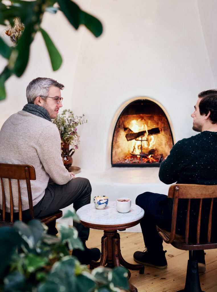 Two people sit by a lit fireplace, talking, with mugs on a small table between them in a cozy room.