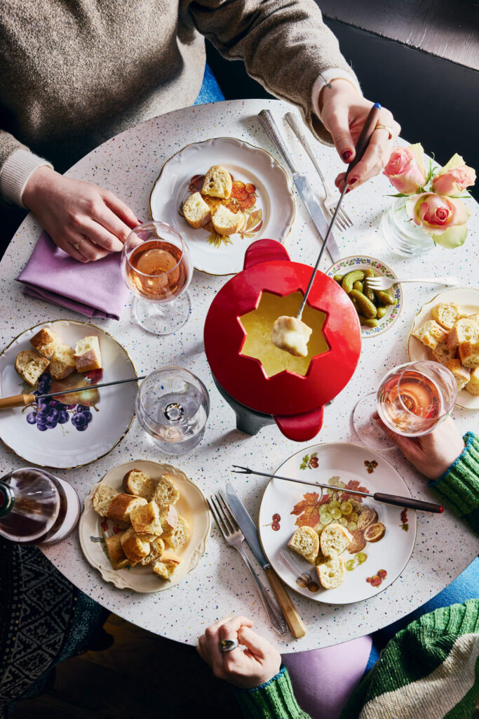 Two people dip bread into cheese fondue at a round table set with plates, wine, and flowers.