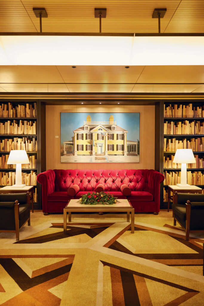 Elegant library with a red velvet sofa, bookshelves, lamps, and a painting of a house above the couch.