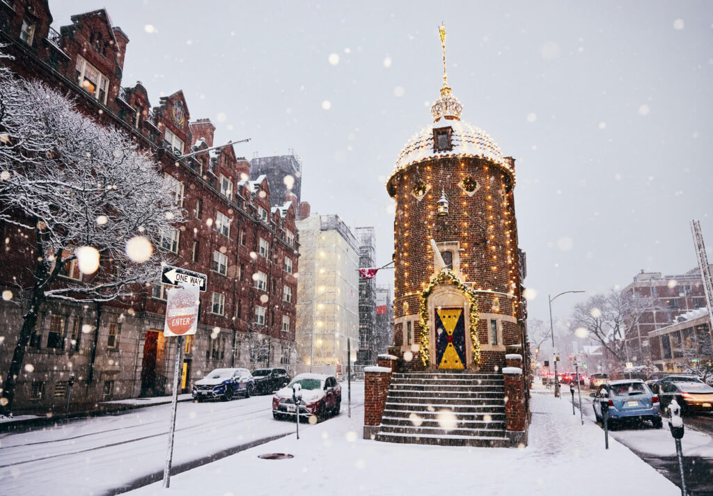 A small, ornate brick tower lit up in the snow on a city street with cars and historic buildings.