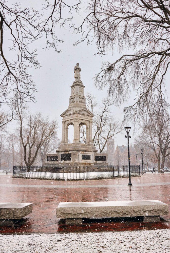 Snow falls on a stone monument surrounded by bare trees in a park with brick pathways.