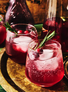 Two glasses of pink sparkling drink with ice, cranberries, and rosemary sprigs on a tray.