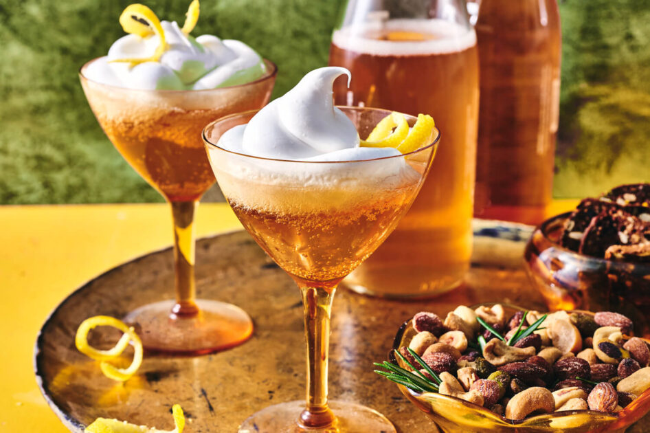 Two cocktails topped with whipped foam and lemon twists, beside a bowl of mixed nuts on a tray.