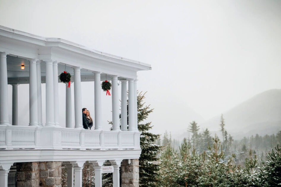 A couple stands on a snowy white-columned porch, surrounded by winter trees and mountains in the background.
