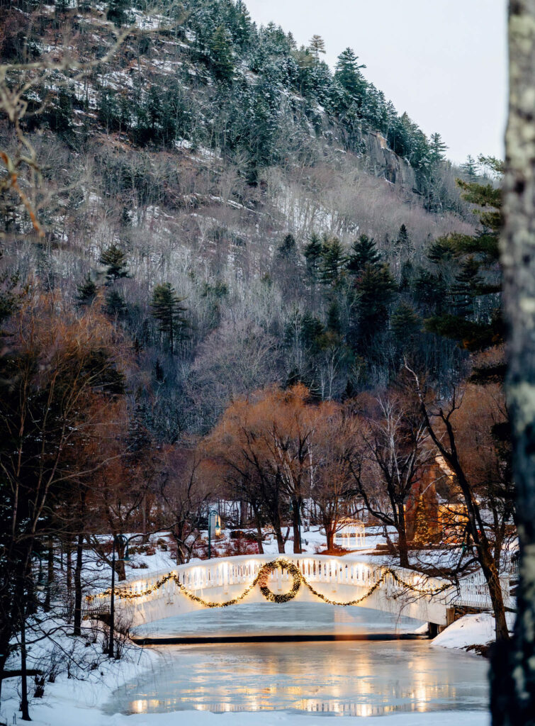 A Christmas Getaway in the White Mountains: Finding Holiday Spirit in New Hampshire. A snowy bridge decorated with lights and a wreath, surrounded by trees and mountains in winter.