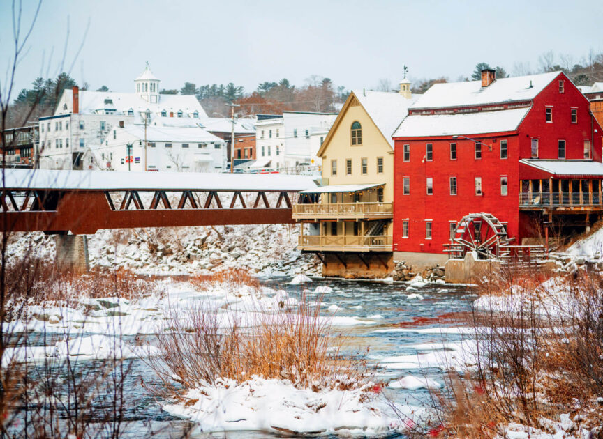 Red mill and covered bridge over a partially frozen river in a snowy small town setting.