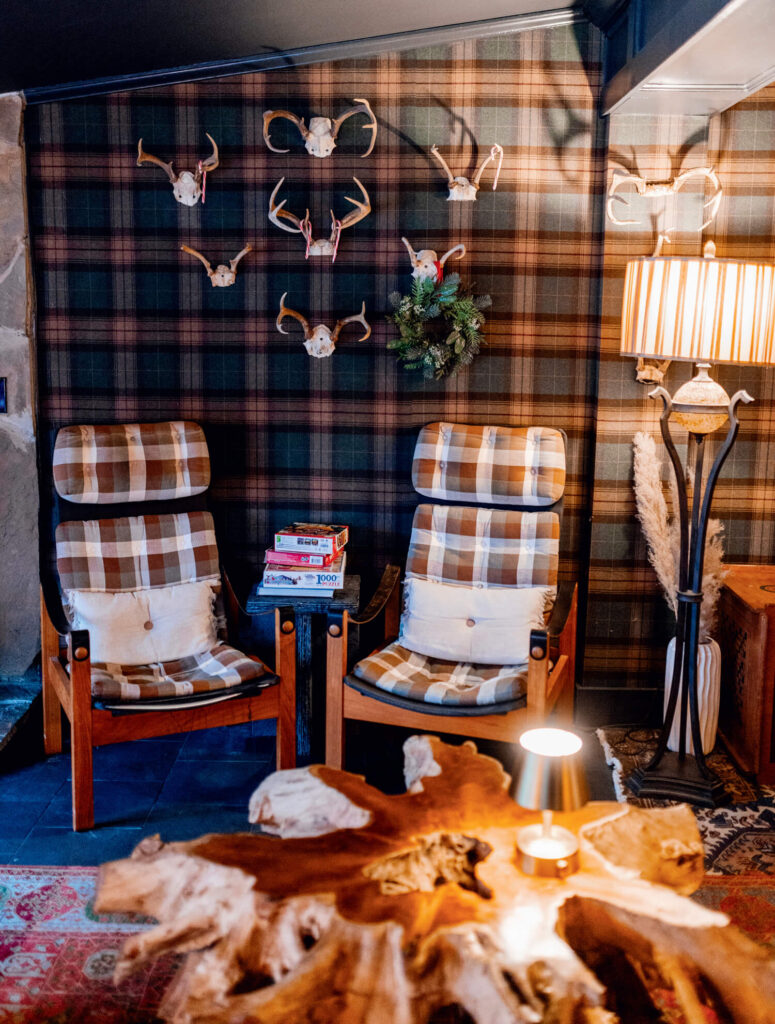 Two plaid armchairs with books, plaid wallpaper, animal skulls, and a floor lamp in a cozy rustic room.