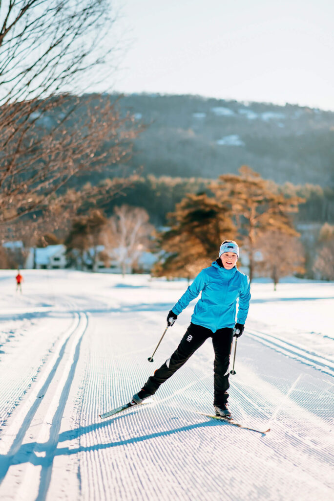 Person cross-country skiing on a snowy trail with trees and mountains in the background.