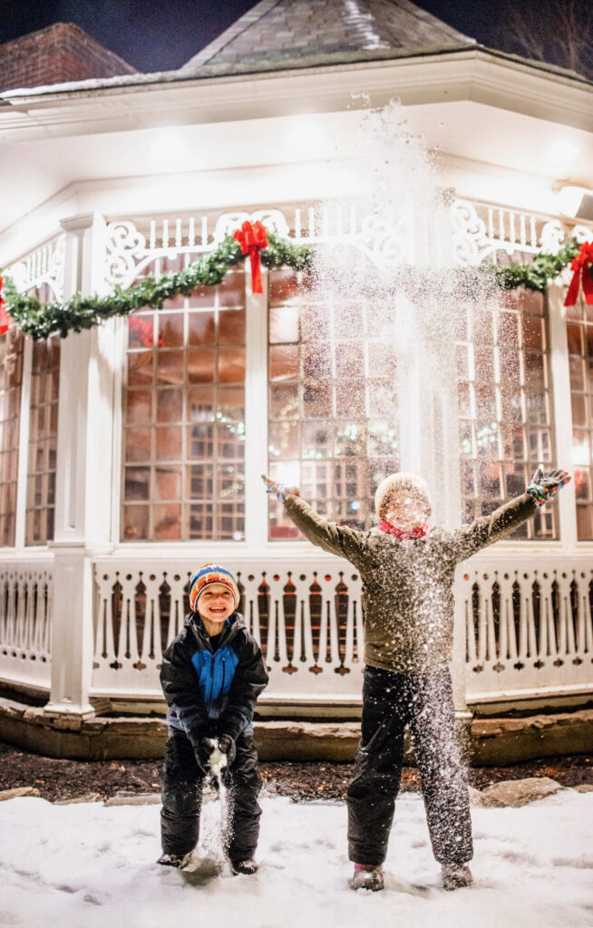 Two children playing in the snow at night in front of a festive, decorated gazebo.