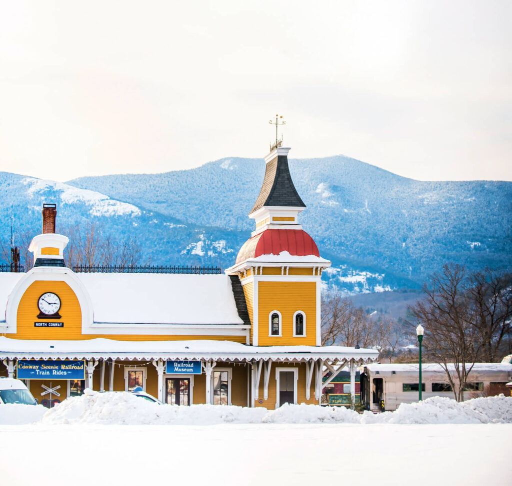 Yellow train station with clock tower, snow-covered roof, and mountains in the background on a winter day.