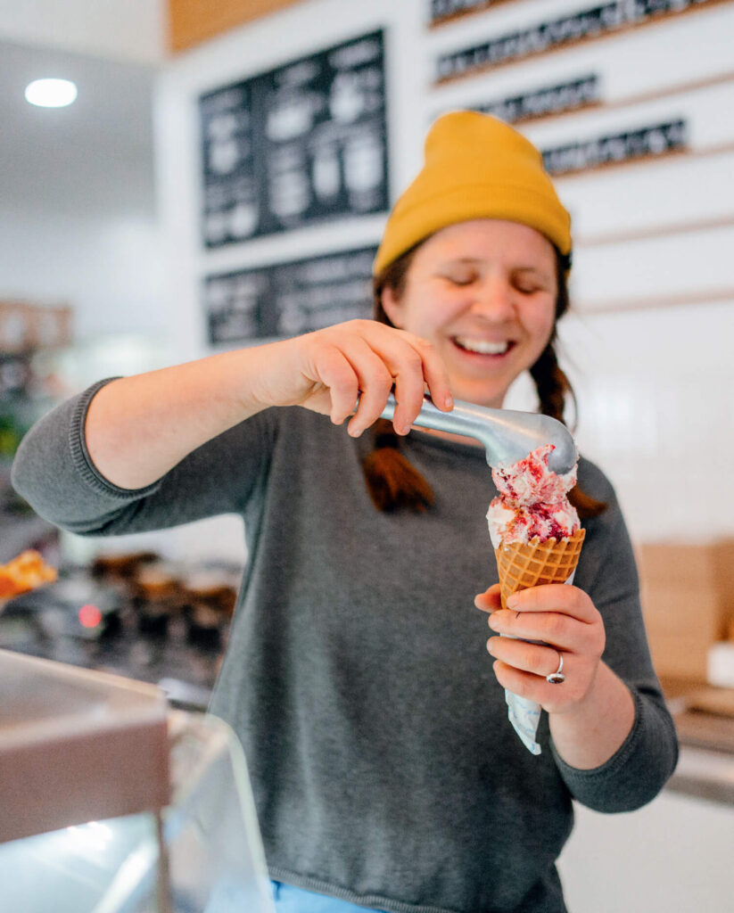 A smiling person in a yellow beanie scoops ice cream into a waffle cone at an ice cream shop.