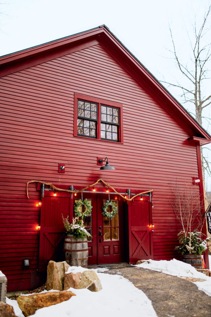 Red barn with double doors, wreaths, and string lights, surrounded by snow and bare trees in winter.