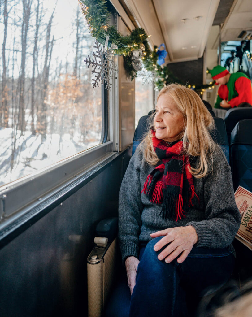 Older woman in a red plaid scarf looks out a train window decorated with garland and snowflakes.