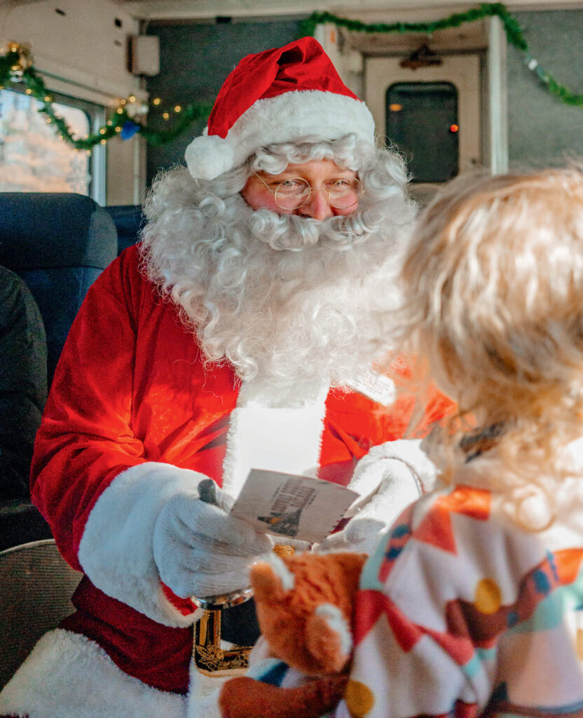 Santa Claus hands a card to a child holding a stuffed toy on a festive, decorated train.
