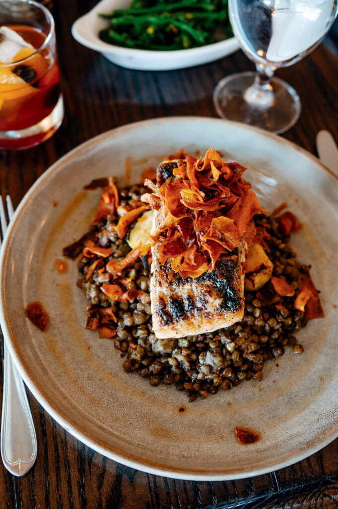 Grilled fish on a bed of lentils, topped with crispy vegetables, served with a drink and greens in the background.