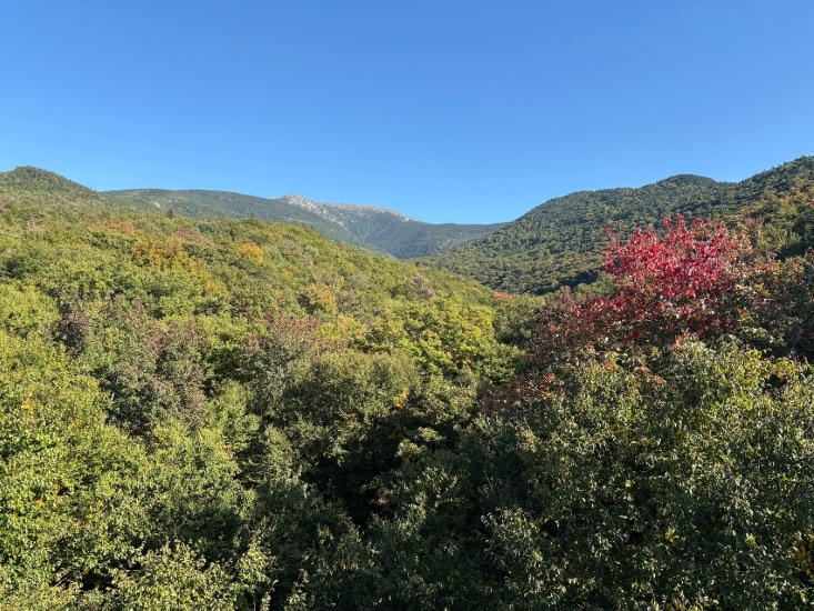 2026 New England foliage map. Green hills with a patch of red foliage under a clear blue sky and mountains in the distance.