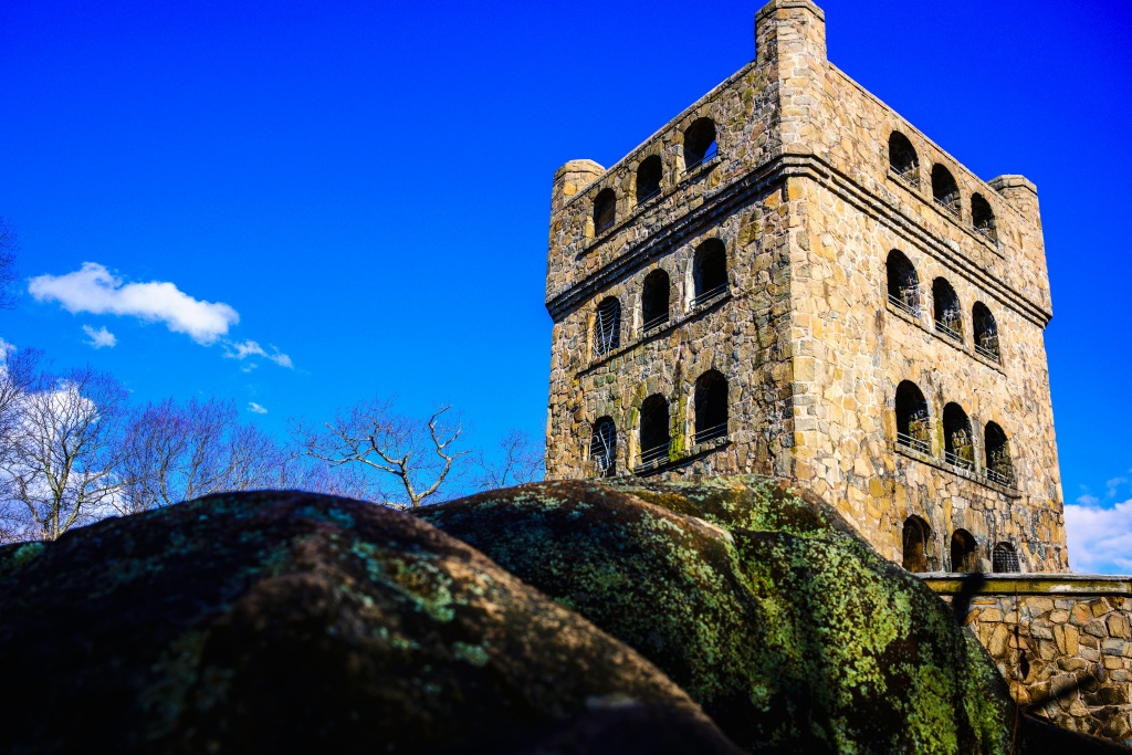 Stone tower with arched windows under a bright blue sky, seen behind large mossy rocks and bare trees.