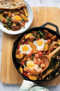 Baked Eggs with Spiced Potatoes. A skillet with eggs, potatoes, veggies, and ham, with a wooden spoon and a plate of the same meal nearby.