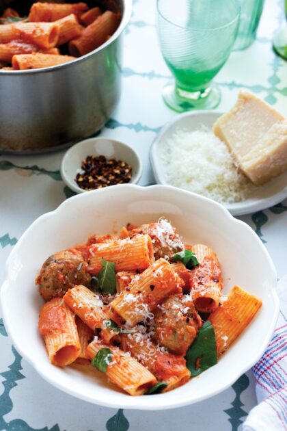 Bowl of rigatoni pasta with meatballs, tomato sauce, basil, and grated cheese, with cheese and pepper flakes nearby.