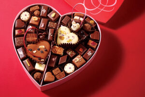 Heart-shaped box filled with assorted chocolates and candies on a red surface.