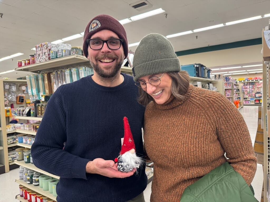 Two people in sweaters and beanies smile in a Renys store in Maine, holding a small gnome figurine.