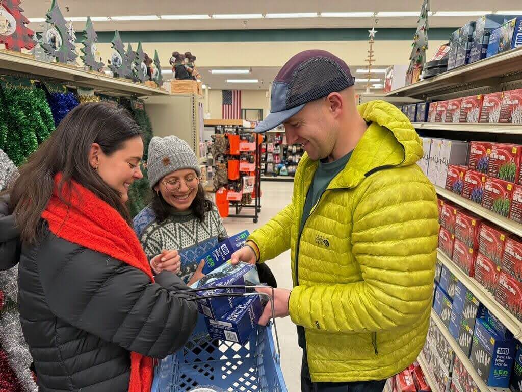 At Renys in Saco, Maine, three people smiling and shopping for holiday lights in a store aisle decorated with Christmas items.