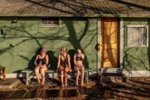 Three women in swimsuits sit on a stone step outside a green wooden building in sunlight.