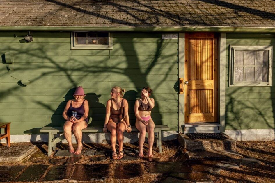 Three women in swimsuits sit on a stone step outside a green wooden building in sunlight.