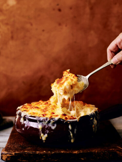 A hand lifts a cheesy serving of baked macaroni and cheese from a brown dish on a wooden board.