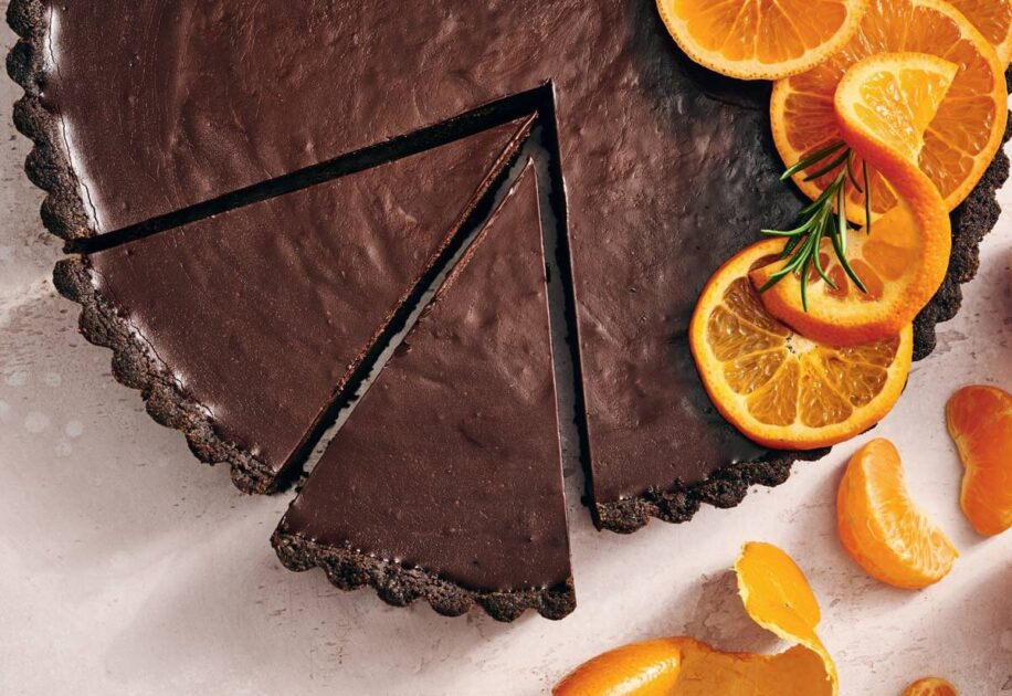 Chocolate tart with a slice removed, garnished with orange slices and rosemary on a light background.
