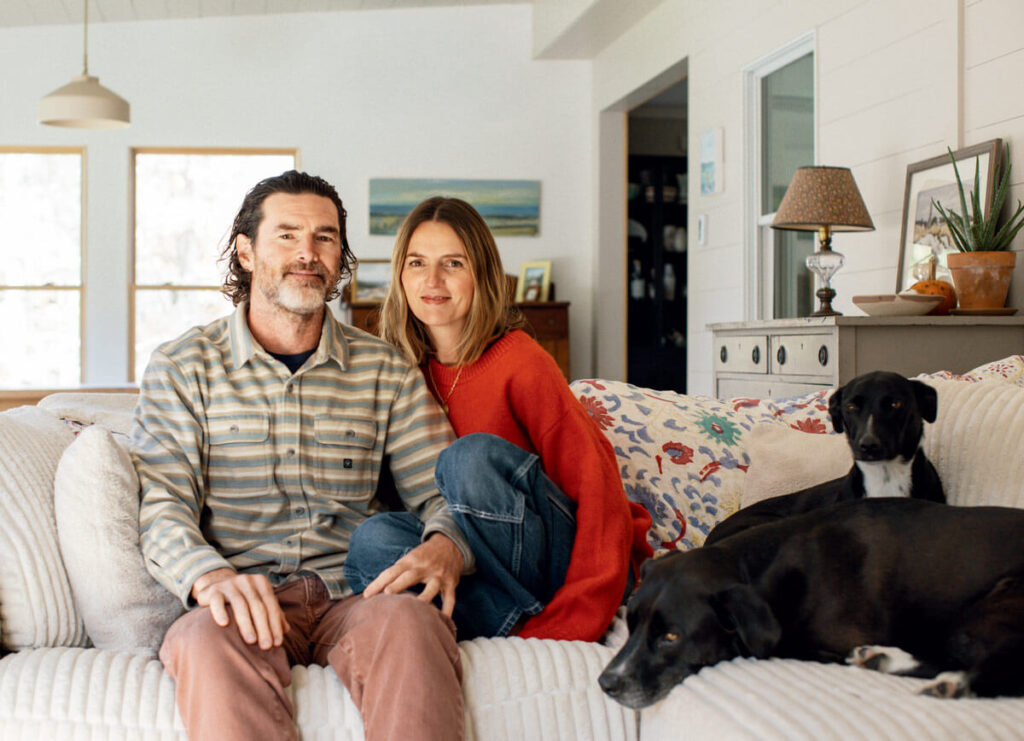 A couple sits on a couch with two black dogs in a cozy, well-lit living room.