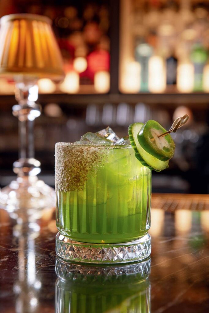 A green cocktail with ice, a salted rim, and sliced jalapeño garnish on a bar counter.