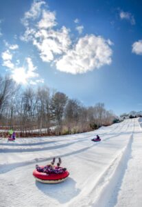 Children slide down a snowy hill in colorful tubes under a bright, sunny sky with scattered clouds.