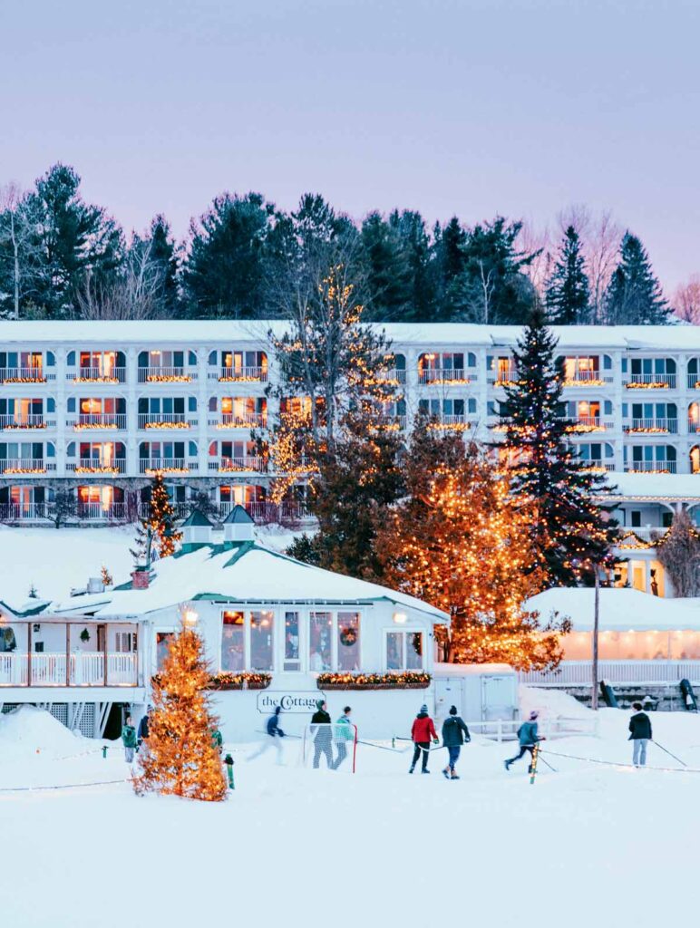 People ice skating near a festive, snowy lodge decorated with Christmas lights at dusk.